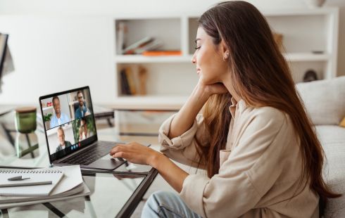 A woman in a home office sits in front of a laptop with a video conference call featuring four other professionals on the screen.