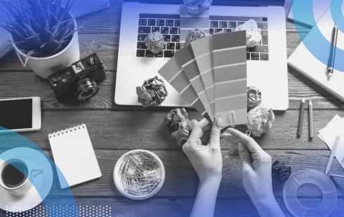 Hands hold color swatches in front of a laptop with a cup of coffee and wadded up balls of paper strewn about