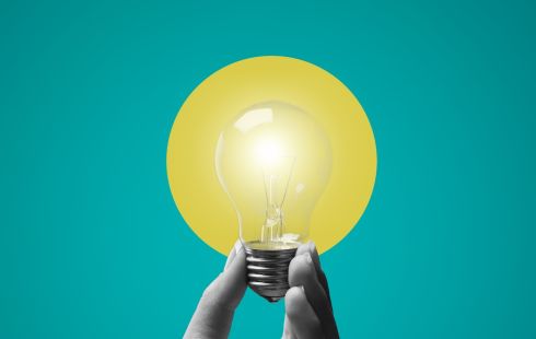 A hand holding a lit light bulb against a blue and yellow background